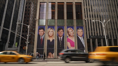 Fox News host photos on billboard on giant building in New York. Cars drive by in front.