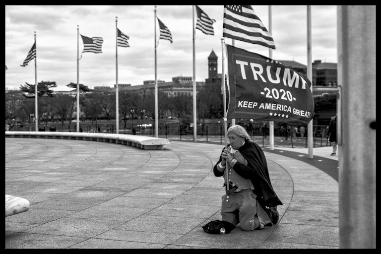 A man in a Revolutionary War costume kneels on the ground, clutching a Trump banner. 