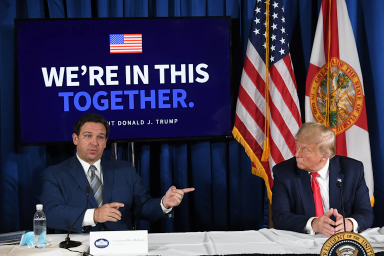 Ron DeSantis and Donald Trump sit at a table with a banner reading "We're in this together" behind them.
