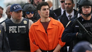 Luigi Mangione in an orange jumpsuit being escorted by law enforcement officials. (A corner of Eric Adams’s face can be seen.)