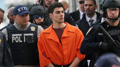Luigi Mangione in an orange jumpsuit being escorted by law enforcement officials. (A corner of Eric Adams’s face can be seen.)