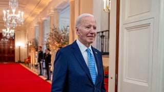 Joe Biden walking. Chandeliers and a red carpet are in the background.