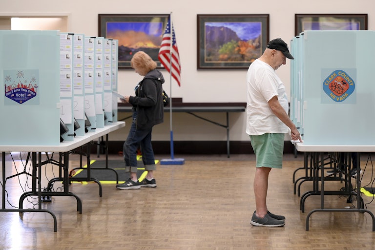 Voters cast their ballots at Desert Vista Community Center in Las Vegas, Nevada. In the foreground to the right, an older white man wearing blue shorts, a white tshirt, and a black cap points at a paper on the table in front of him. In the background to the left, a woman with strawberry blonde hair wearing jeans and a black hoodie looks at a ballot. The booth panel has a sticker that reads "Clark County, Nevada."