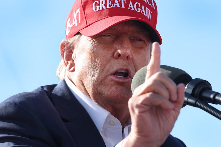 Donald Trump wearing a red Make America Great Again cap speaks into a mic and holds up his right index finger. He is squinting.