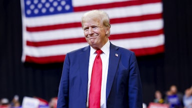Donald Trump toothlessly smiles as he stands before a large American flag at a political rally