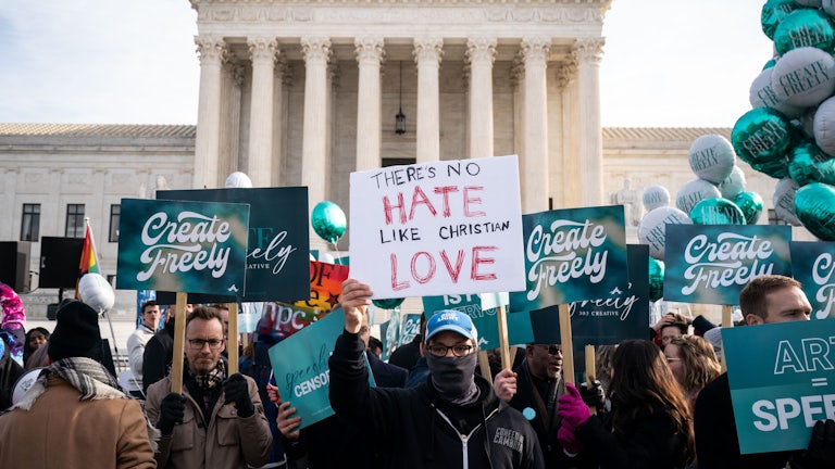 A crowd of protesters gather in front of the Supreme Court building. One protester holds up a sign that says THERE'S NO HATE LIKE CHRISTIAN LOVE, while others hold up signs that say CREATE FREELY.