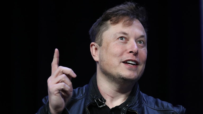 Elon Musk smiles, points a finger upwards 