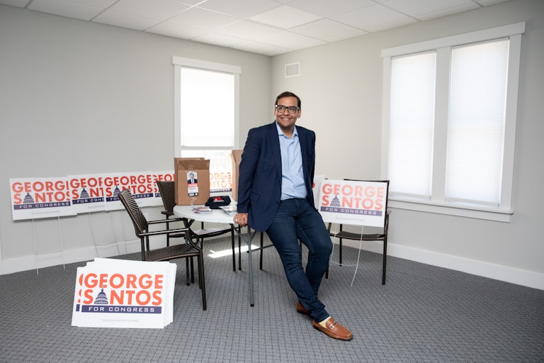 George Santos leans on a table in what looks like an empty office space. He smiles and looks to his right. Around him are several "George Santos for Congress" yard signs.