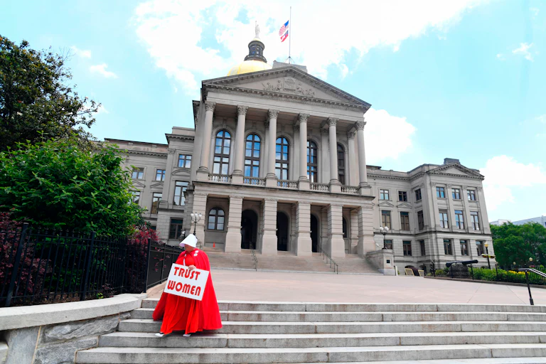 A woman wearing an outfit from The Handmaid's tale stands on the steps of the Georgia Capitol, holding a sign that reads "Trust Women."