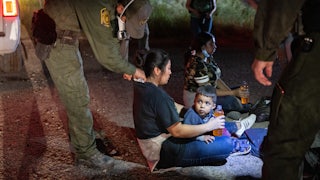 A Border Patrol agent bends down to place something on the neck of an undocumented immigrant sitting on the ground with a child in her lap.