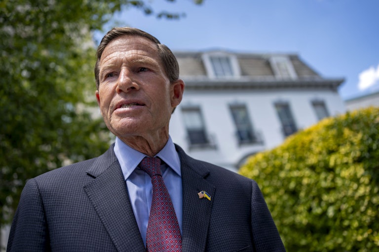 Richard Blumenthal stands outside, in front of some bushes and a house.