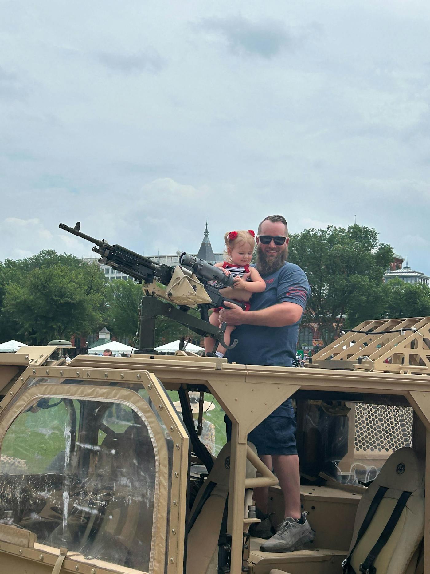 A white man with a large bears smiles as he props up his toddler daughter in front of a massive gun at Trump's military parade.