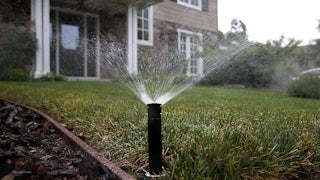 A sprinkler sprays water over a lawn with a house in the background.