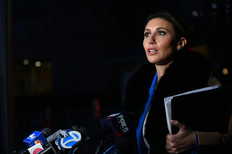 Alina Habba looks surprised. She is standing outside at nighttime and several press mics stand before her. She holds a folder of papers in her left hand.
