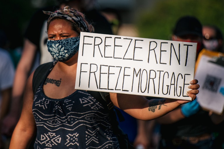 A woman at a protest holds a sign that reads "Freeze Rent, Freeze Mortgage." 