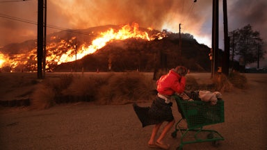 A person pushes a cart of belongings with a burning hill in the background.