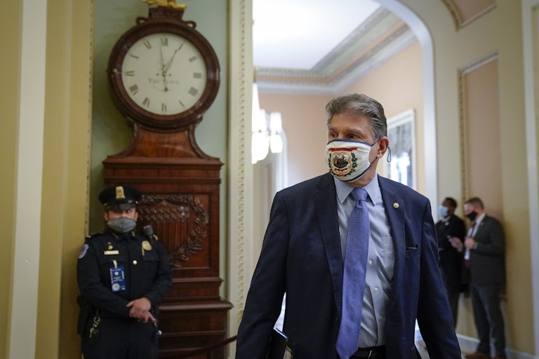 Clad in a mask with the West Virginia state flag on it, Senator Joe Manchin walks down a Capitol hallway in front of a masked guard.