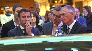 Carlos Tavares looks at Emanuel Macron, who frowns at a car.