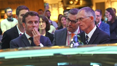 Carlos Tavares looks at Emanuel Macron, who frowns at a car.