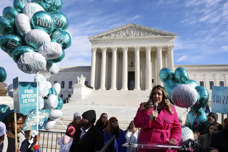  A woman in a pink coat speaks into a microphone at a podium in front of the Supreme Court. She is surrounded by supporters carrying signs and balloons that say CREATE FREELY and FREE SPEECH IS FOR EVERYONE.