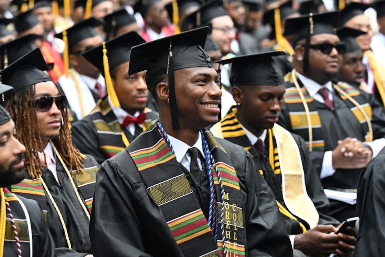 Morehouse College graduates wearing caps and gowns participate in the commencement ceremony. A student smiles in the foreground.