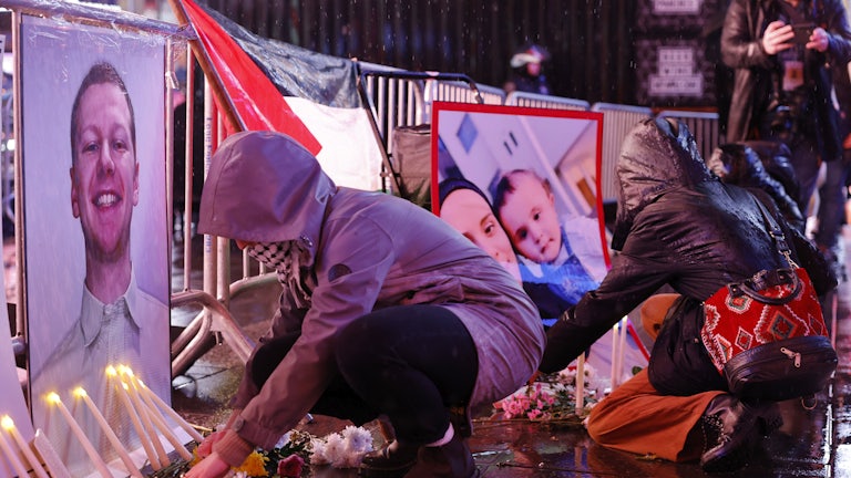 People in rain jackets light candles and crouch in front of pictures and flowers.