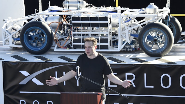 Elon Musk stands at a podium in front of a vehicle on a platform with a banner underneath reading HYPERLOOP POD COMPETITION.