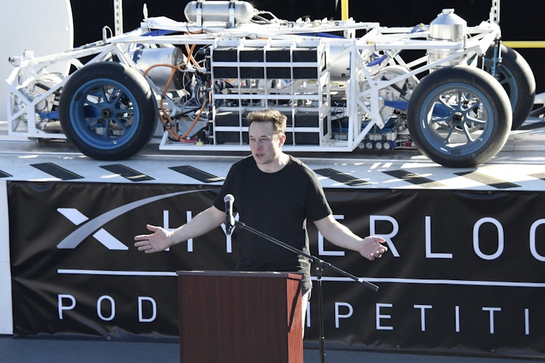 Elon Musk stands at a podium in front of a vehicle on a platform with a banner underneath reading HYPERLOOP POD COMPETITION.