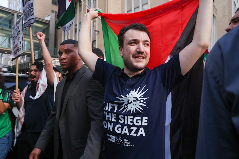 Mahmoud Khalil wears a shirt that reads "Lift the Siege on Gaza" and carries a Palestinian flag with both hands.