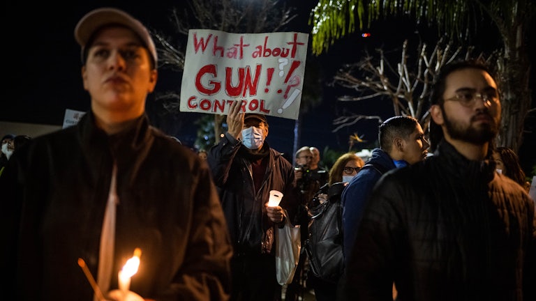 Someone wearing a mask holds a sign that reads "What about gun control?" and a candle. Others around him also hold candles.
