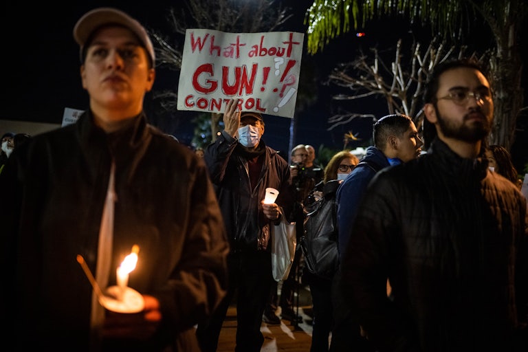 Someone wearing a mask holds a sign that reads "What about gun control?" and a candle. Others around him also hold candles.