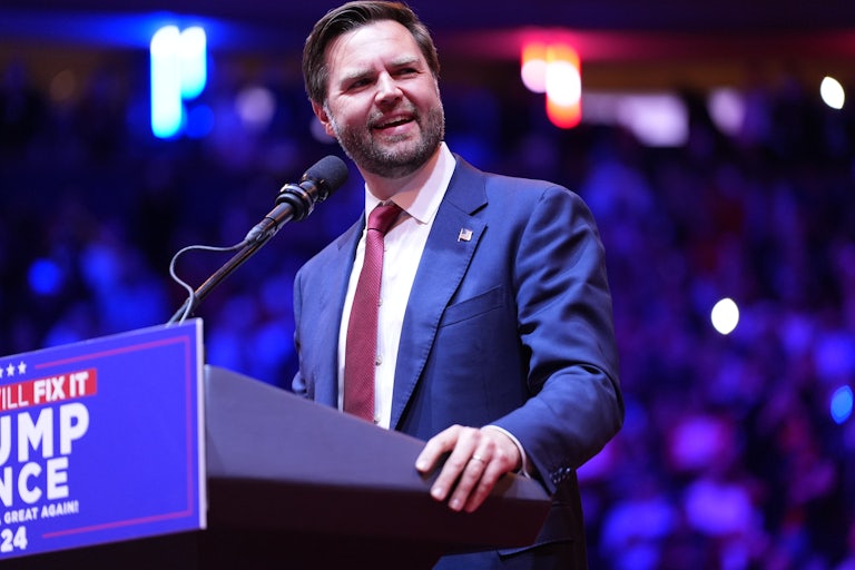 JD Vance smiles and looks to the side while standing at a podium during a Donald Trump rally