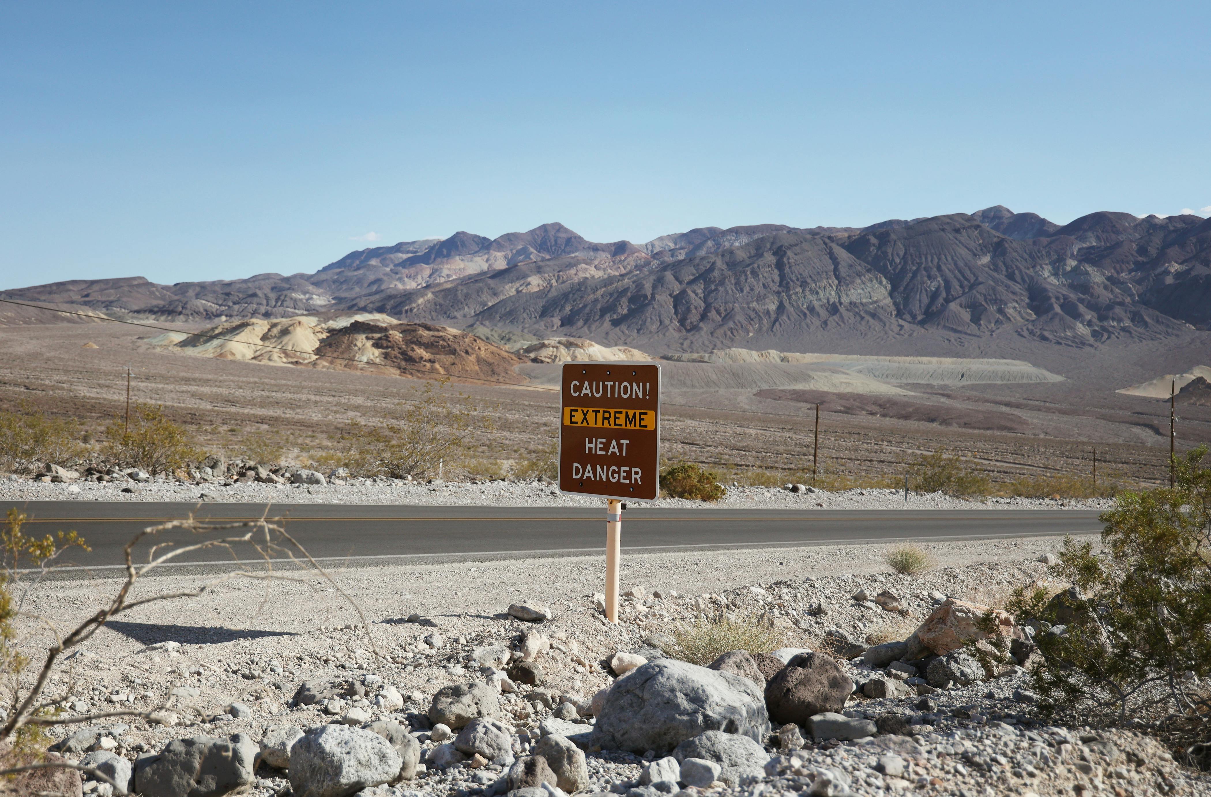 A desert road, devoid of cars, shows a sign that says CAUTION! EXTREME HEAT DANGER. The sun is out and there are mountains in the background.