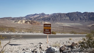 A desert road, devoid of cars, shows a sign that says CAUTION! EXTREME HEAT DANGER. The sun is out and there are mountains in the background.