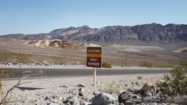 A desert road, devoid of cars, shows a sign that says CAUTION! EXTREME HEAT DANGER. The sun is out and there are mountains in the background.