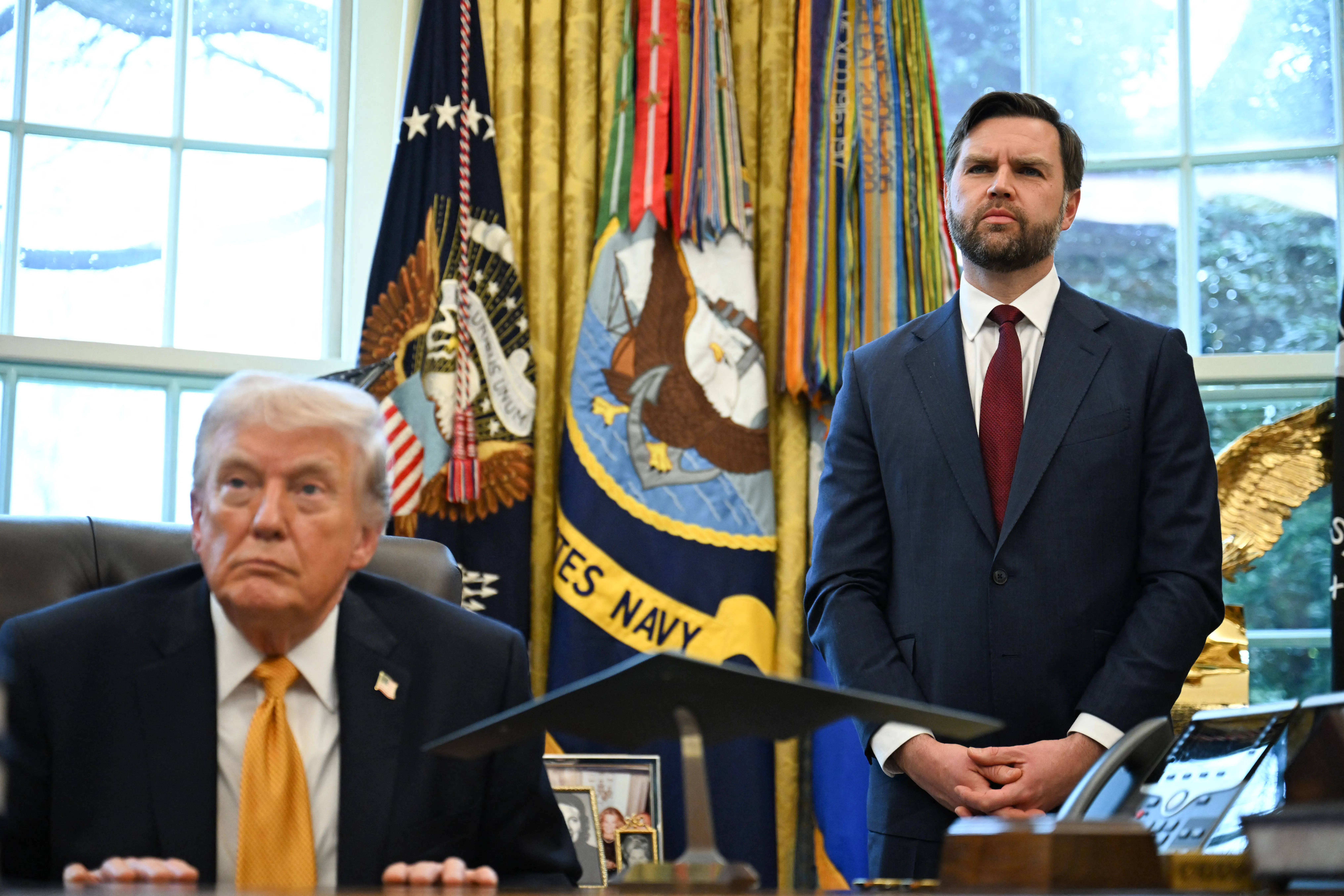 Donald Trump sits at his desk in the Oval Office of the White House as JD Vance stands next to him