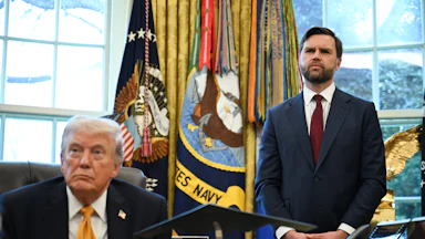 Donald Trump sits at his desk in the Oval Office of the White House as JD Vance stands next to him