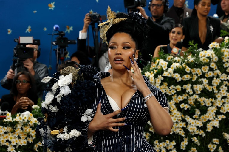 Nicki Minaj poses for the cameras on a red carpet. She rests her super long fingernails on her face and chest.