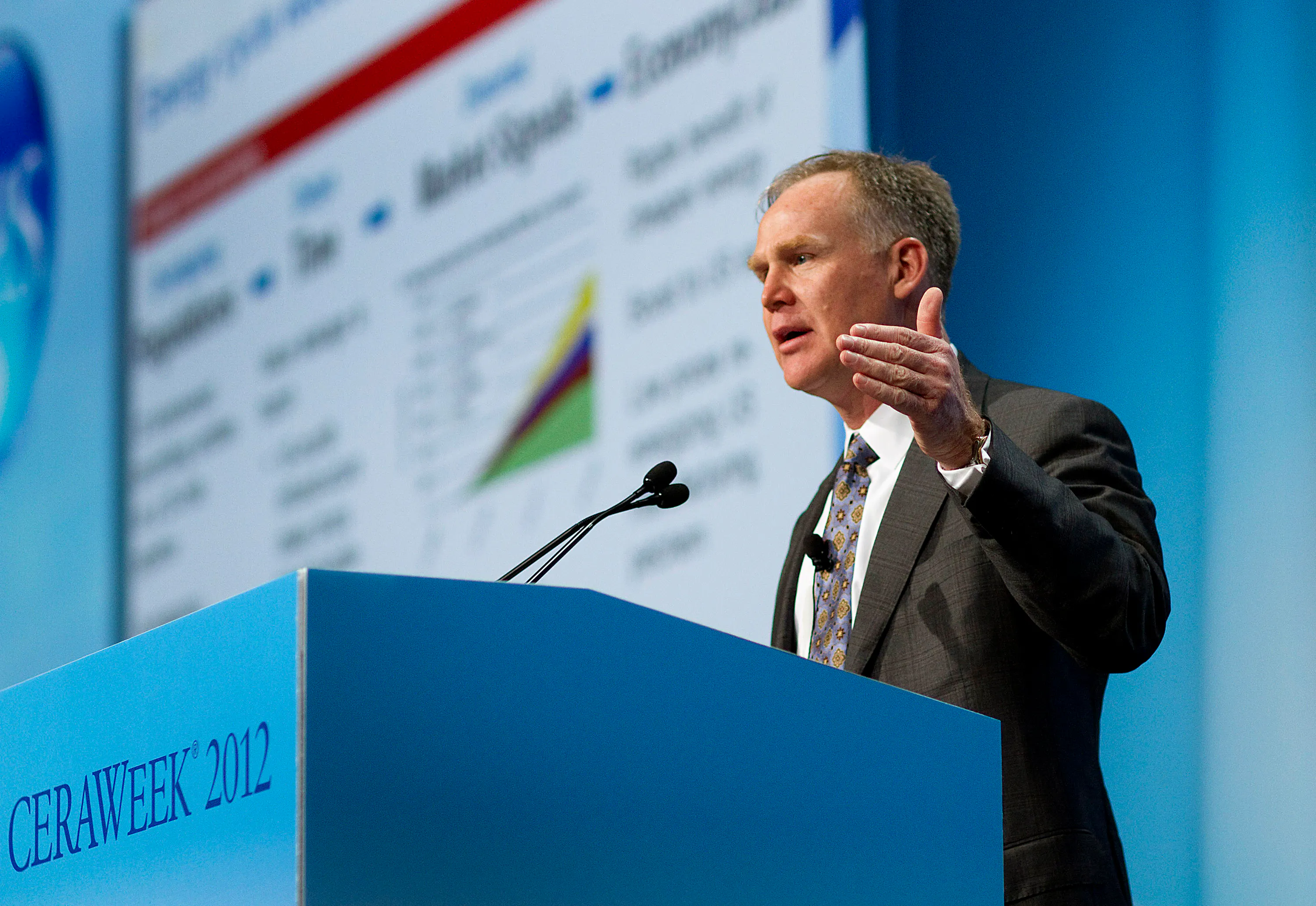 Alan Armstrong speaks behind a blue podium with CERAWEEK 2012 written on the front and a faint presentation behind him to his right.