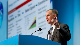 Alan Armstrong speaks behind a blue podium with CERAWEEK 2012 written on the front and a faint presentation behind him to his right.