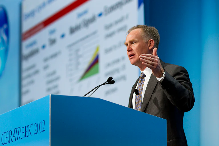 Alan Armstrong speaks behind a blue podium with CERAWEEK 2012 written on the front and a faint presentation behind him to his right.