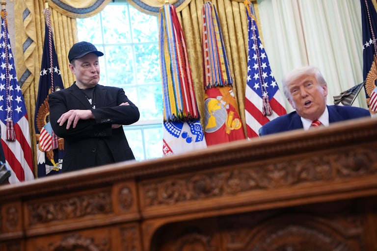 Elon Musk purses his lips and crosses his arms while standing next to Donald Trump, who speaks while sitting at his desk in the Oval Office