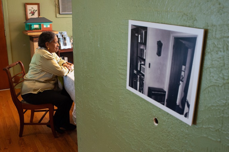 Myrlie Evers-Williams sits at a table in the Medgar and Myrlie Evers Home National Monument. A black and white photo is on the wall.