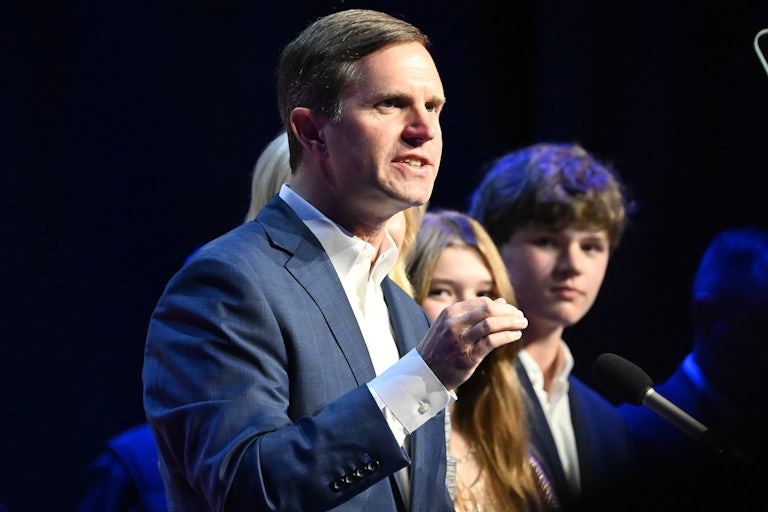 Andy Beshear speaks before a mici and makes a hand gesture. A couple kids are in the background.