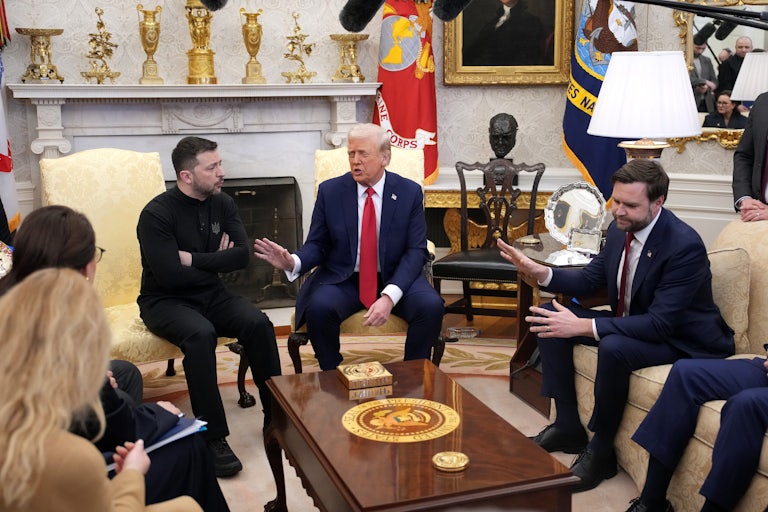 Ukraine's President Volodomyr Zelenskiy, Donald Trump, and J.D. Vance sit in the White House along with membres of the press and select visitors. Zelenskiy has his arms crossed and is speaking, while both Trump and J.D. Vance hold a hand up as if to rebuff what he is saying.