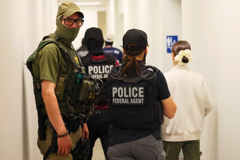 A masked immigration agent looks directly at the camera as two other people in police vests steer a woman away.