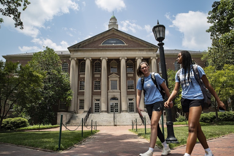 Two female students (one white, one black) walk in front of a building at the University of North Carolina Chapel Hill.
