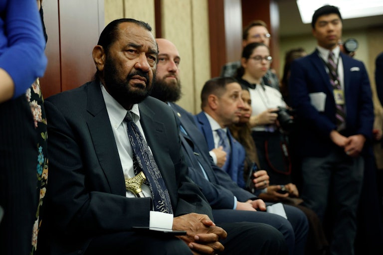 Representative Al Green sits on a chair and listens to someone while his hands are clasped. Several people sit and stand around him, also listenin.