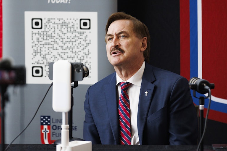 Mike Lindell speaks in a mic, his brows furrowed. A giant QR code is behind him.