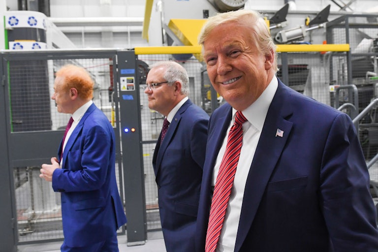 Donald Trump smiles in the foreground. Anthony Pratt and Scott Morrison are in the background. The three men are walking around a plant.